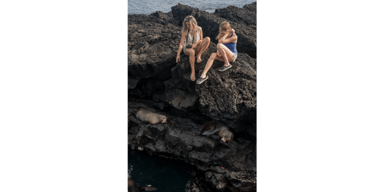 Two tourists watching two Galapagos fur seals resting in Santiago Egas Port